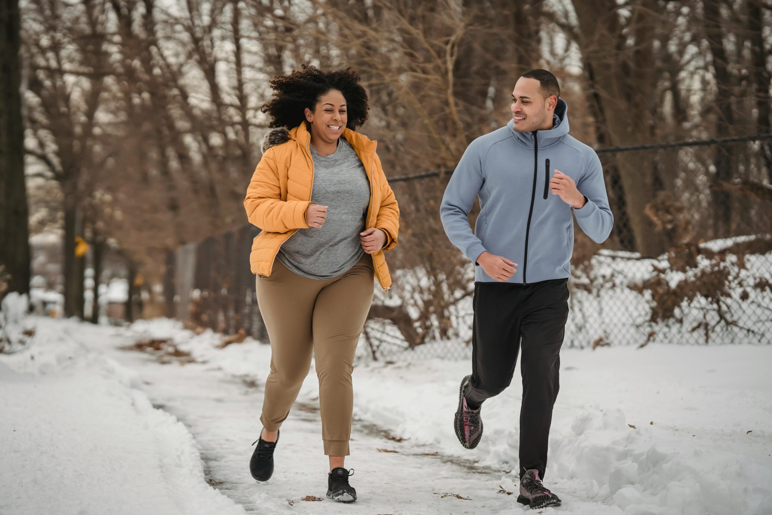 Full length smiling ethnic male fitness trainer and plus sized black female in warm jacket jogging together on snowy pathway in frozen winter park