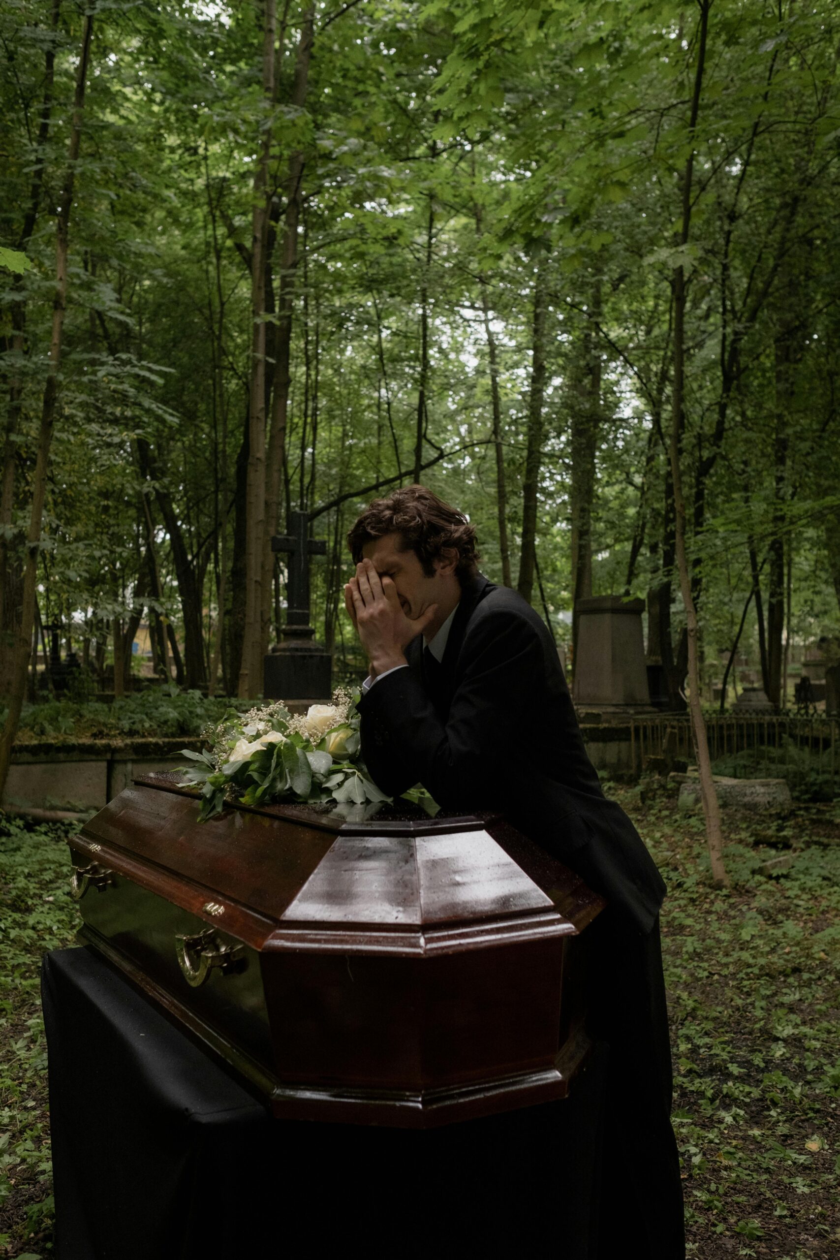 A man grieving at a funeral, leaning on a flower-adorned coffin in a forested cemetery.