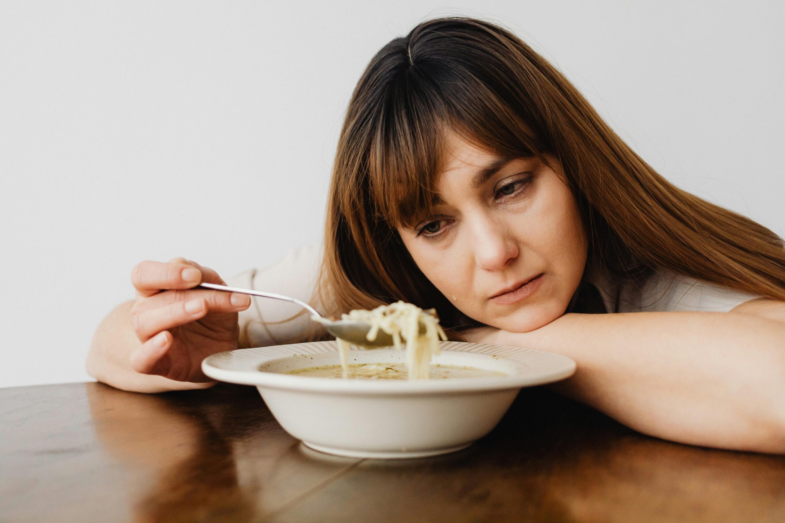 A woman sitting at a table thoughtfully gazing at a bowl of noodle soup, evoking a sense of contemplation.