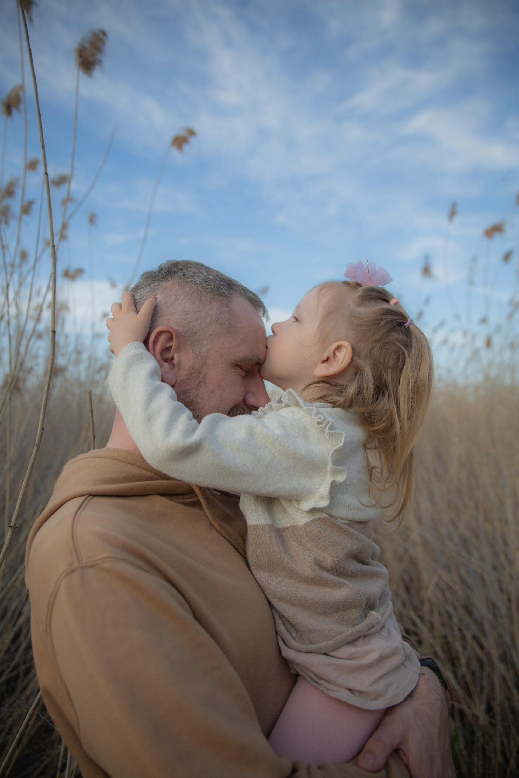 A tender moment with a father and daughter embracing in a grassy field under a blue sky.