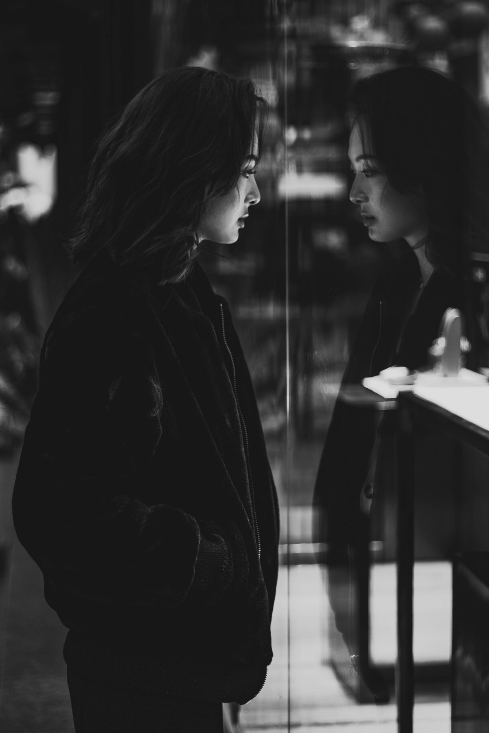 Black and white portrait of an Asian woman reflecting in glass, capturing her thoughtful expression.
