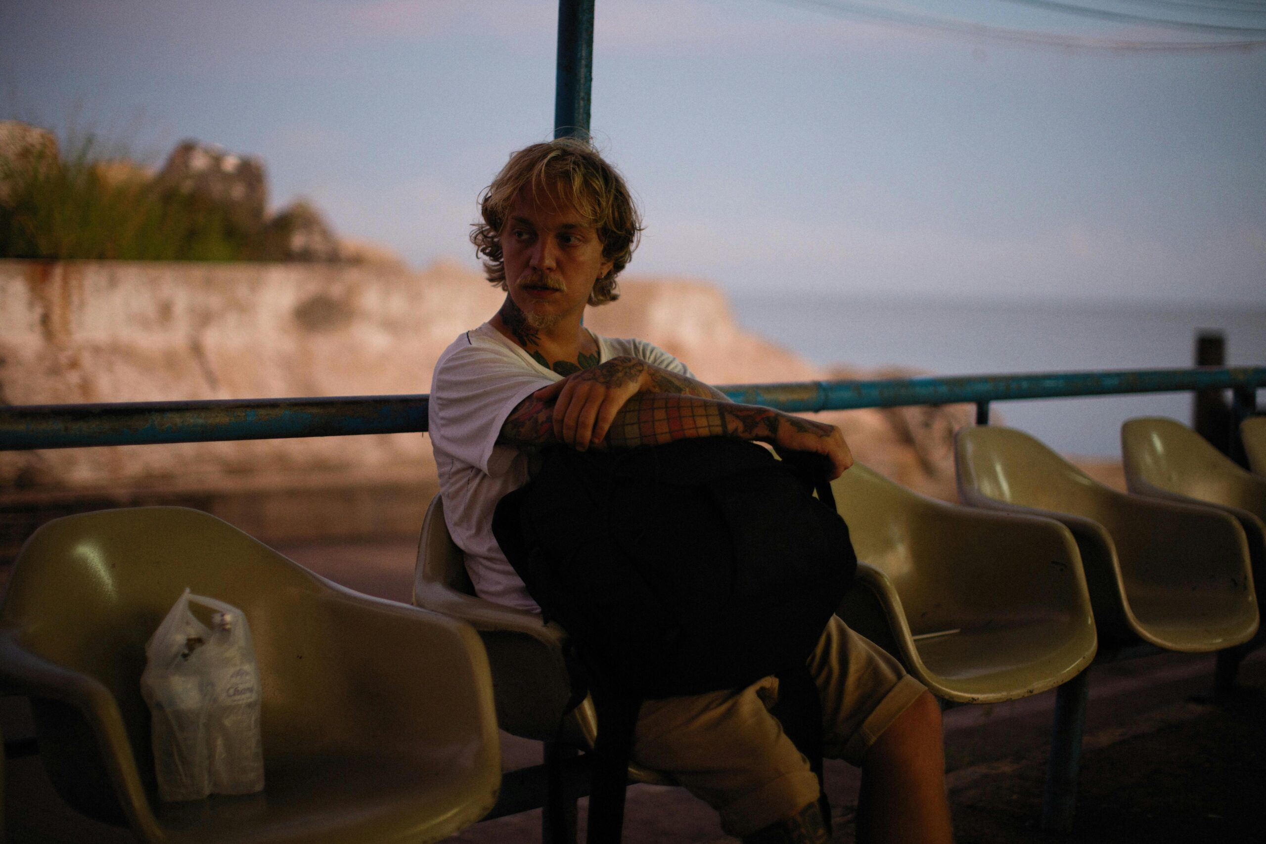 A man sits thoughtfully on an outdoor chair, holding a bag, during twilight.